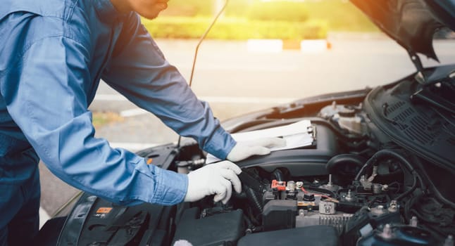 Close-up of a professional Asian mechanic's hand, in blue workwear, writing on a paper while working on a car in an auto repair shop.