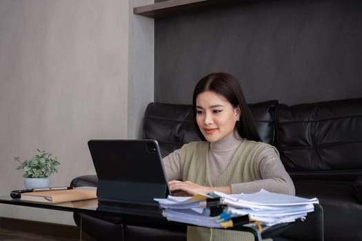 A middle-aged woman works from home at the dining room table with a laptop and a stack of documents next to her..