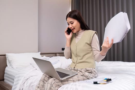 A middle-aged woman works from home at the bed room with a laptop and a stack of documents next to her..