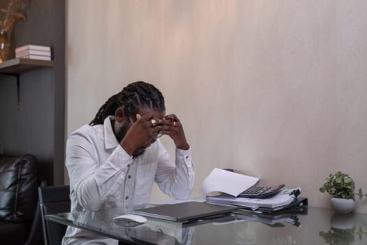 Young African American businessman feeling stressed and tired while working from home with documents stacked next to him..