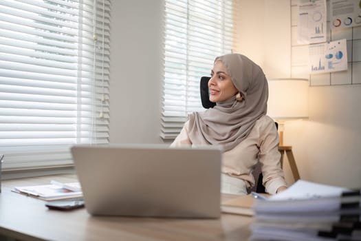 Muslim female entrepreneur wearing hijab sits working with laptop managing personal business in private office..