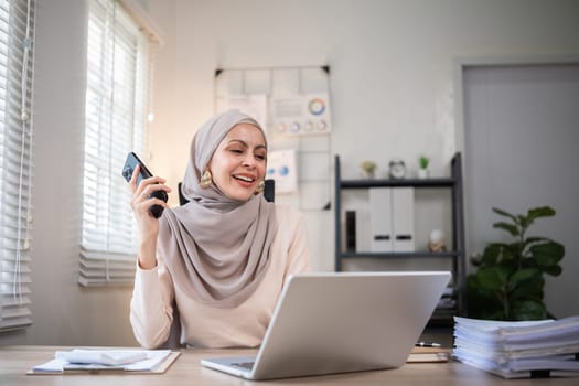 Muslim female entrepreneur wearing hijab sits working with laptop managing personal business in private office..