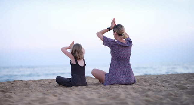 Mother and daughter do yoga meditate in lotus position on beach. Family yoga class by sea concept