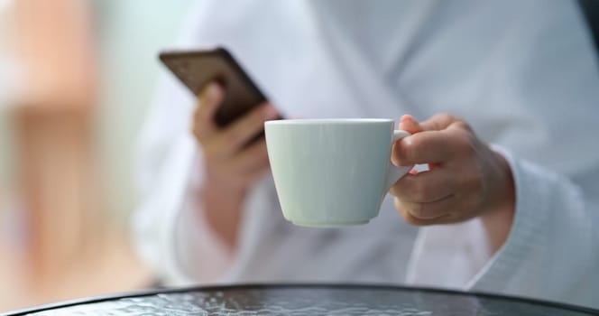 Cropped photo of a woman in a terry bathrobe holding mobile phone and cup. Watching news and relaxing morning coffee