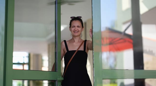 Beautiful smiling young woman opens glass door. Satisfied customer or meeting guests concept