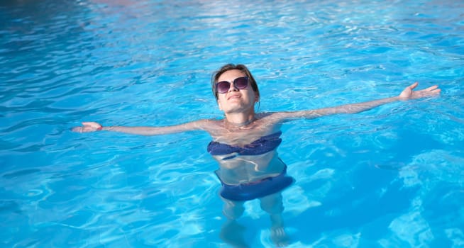 Charming woman in sunglasses enjoys swimming in turquoise water of pool. Relax in luxury hotel pool and wellness concept