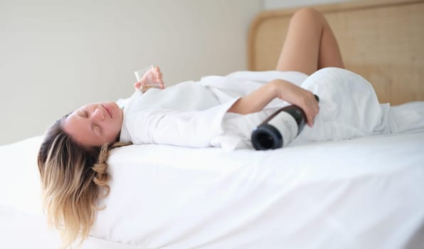Drunk woman in bathrobe sleeps on bed with bottle of wine and glass in hotel room. Female alcoholism stress life problems or drunk female tourist on vacation