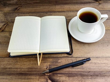 Coffee in a cup and a notebook with a pen on a wooden table. Coffee in a cup with a saucer and a notebook with a pen on a wooden table.