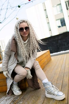 A woman sitting on a simple wooden bench, with a pair of sneakers on her feet. She appears relaxed and casual in her attire, enjoying a moment of rest outdoors.