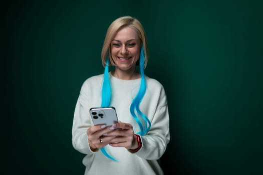 A woman with vibrant blue hair is seen holding a cell phone in her hand. She appears to be looking at the screen intently, possibly texting or browsing. The background is blurred, focusing on the woman and her phone.