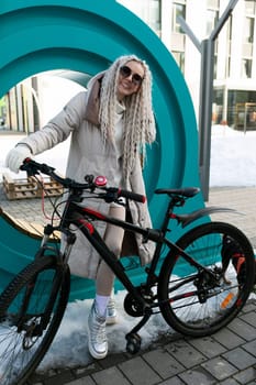 A woman is standing next to a bike parked on a sidewalk in an urban setting. She appears to be waiting or taking a break while holding onto the handlebars of the bicycle.