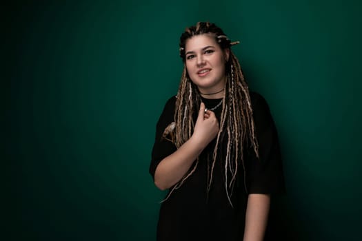 A woman with dreadlocks is standing in front of a green wall. She is dressed casually and looking straight ahead. The background is a solid green color, providing a simple yet vibrant backdrop.