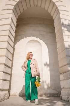 A woman is standing in front of a building with a yellow flower in her hand. She is wearing a green dress and a tan coat