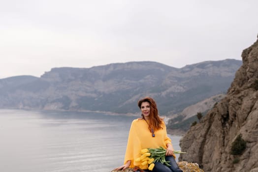 Portrait happy woman woman with long hair against a background of mountains and sea. Holding a bouquet of yellow tulips in her hands, wearing a yellow sweater.