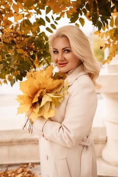 A blonde woman is holding a bouquet of yellow leaves. She is smiling and she is happy