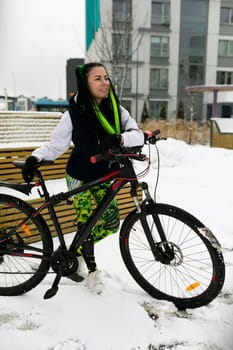 A woman in casual attire sits on a wooden bench next to a bicycle. She appears relaxed, looking towards the distance. The bike leans against the bench, under the shade of a tree.