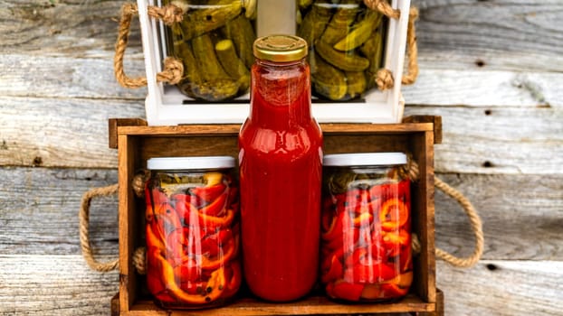 Wooden crate with bottles with tomatoes sauce and glass jars with pickled red bell peppers isolated in a rustic composition. Jars with variety of pickled vegetables preserved food concept.