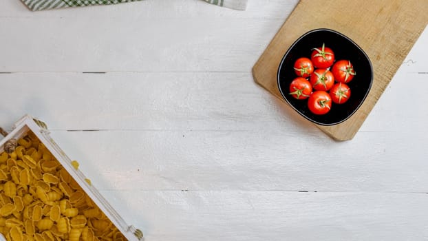 Top view of pasta in a wooden crate and fresh ripe cherry tomatoes in a black bowl on a rustic white wooden table. Ingredients and food concept