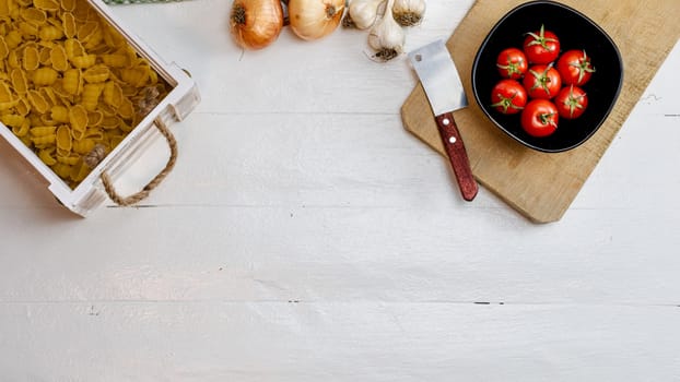 Top view of onions, garlic, pasta in a wooden crate and fresh ripe cherry tomatoes on a rustic white wooden table. Ingredients and food concept
