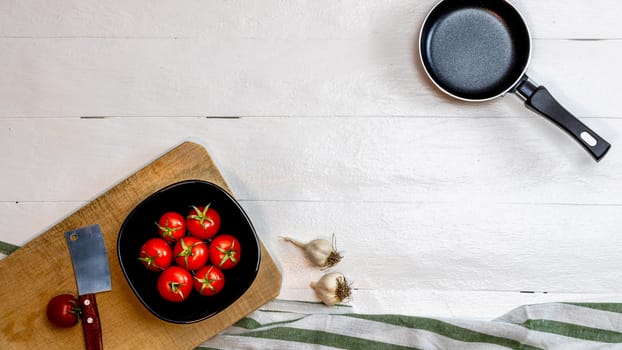 Top view of knife, small pan, onions, garlic and fresh ripe cherry tomatoes in small black bowl on a rustic white wooden table. Ingredients and food concept