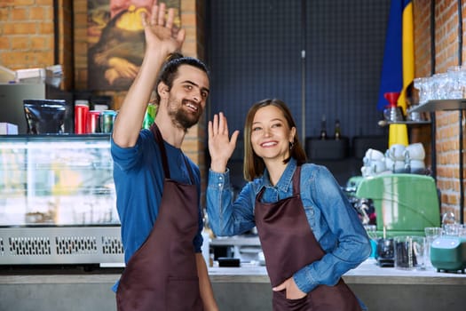 Business team, portrait of confident successful colleagues partners happy cheerful young man and woman in aprons waving hand welcoming guests, at workplace in restaurant coffee shop cafeteria