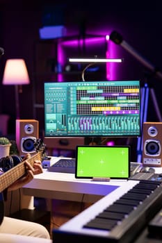 Audio producer practices playing guitar using learning tutorial on tablet via greenscreen display. Young artist learning to play acoustic instrument with online lessons, recording tunes.