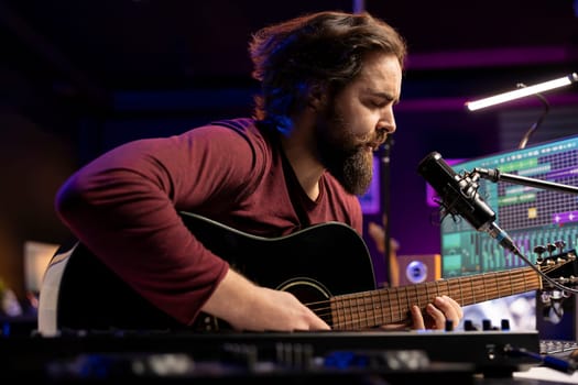 Songwriter composing a new song on acoustic guitar in home studio, using a microphone to sing the chords. Artist works with mixing console and daw software, guitarist producing new music.