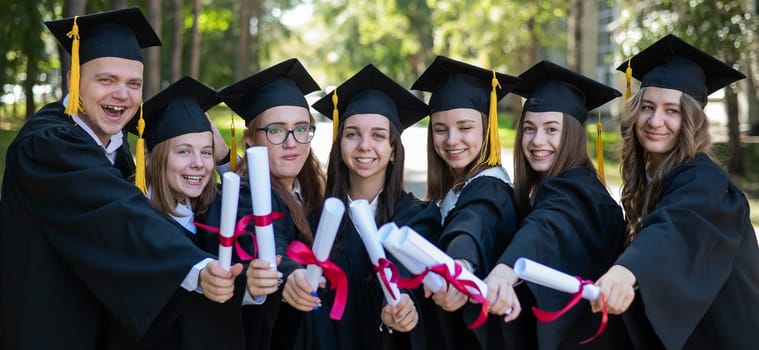 Row of happy young people in graduation gowns holding diplomas outdoors