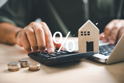 Hand on calculators, strategizing home refinance. Wooden house model, buy and rent note on desk. Smart money management for buying property concept. Tax, analysis for mortgage payment.