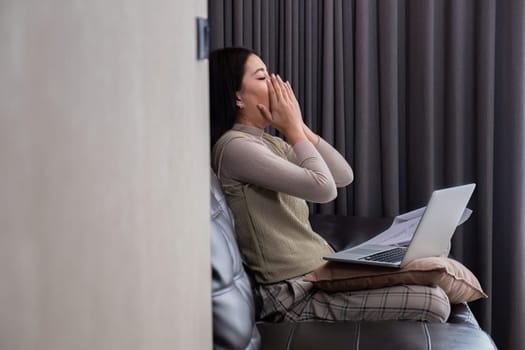 A middle-aged woman is tired while working from home in her bedroom with her laptop and pile of documents next to her..