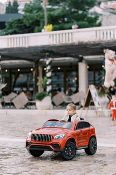Little girl rides a red toy car across the square in front of the building. High quality photo