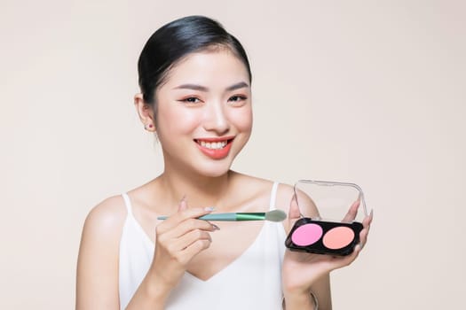 Young asian smiling woman holding blush palette with a brush isolated on a beige background
