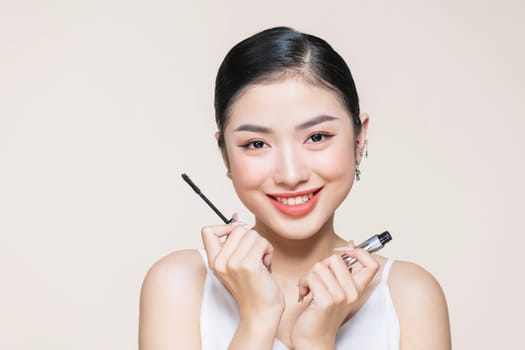 Beautiful asian young woman holding mascara and smiling on purple studio background,