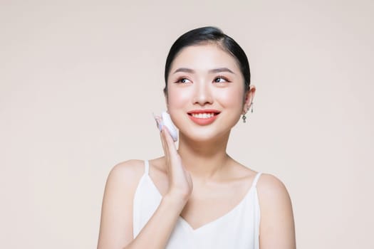 Young asian woman smile looking happy applying makeup with puff makeup on beige background.