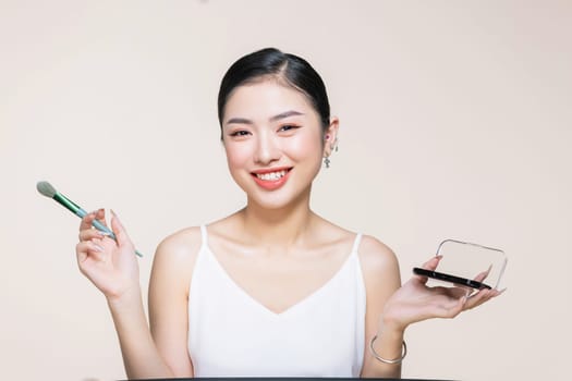 Young asian woman doing makeup with brush while standing on beige background