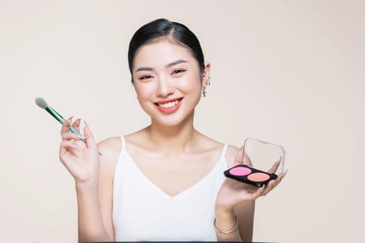 Young asian smiling woman holding blush palette with a brush isolated on a beige background