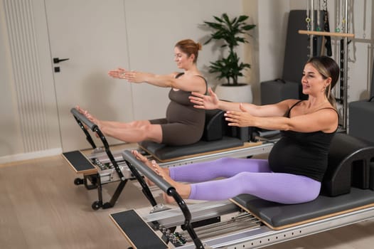Two pregnant women do Pilates exercises on a reformer. Yoga class for pregnant women