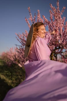 Woman blooming peach orchard. Against the backdrop of a picturesque peach orchard, a woman in a long pink dress and hat enjoys a peaceful walk in the park, surrounded by the beauty of nature