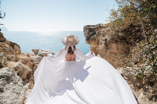 A woman in a white dress is standing on a rocky beach with her hat on. The scene is serene and peaceful, with the woman enjoying the view of the water.