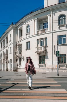 Woman city road crossing. Stylish woman in a hat crosses the road at a pedestrian crossing in the city. Dressed in white trousers and a jacket with a bag in her hands