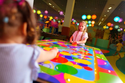 A joyful young girl is captured engaging in a playful board game, surrounded by bright and whimsical decorations in a colorful playroom setting. A hand is seen reaching for a game piece on the reflective, rainbow-hued surface, emphasizing the dynamic and interactive aspects of the game.