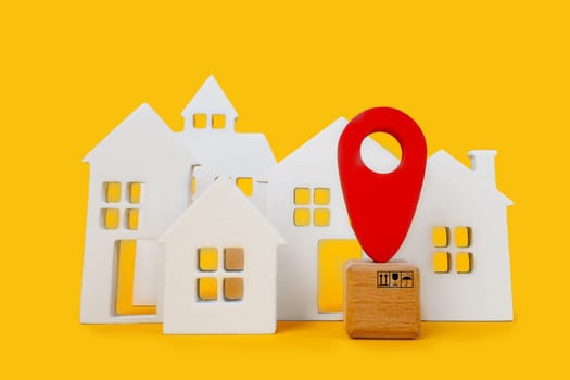 Cardboard boxes and a red position pin in city of white toy house on yellow background. Locating packages and goods.