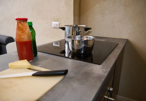 Still life. A stainless steel saucepan on the surface of electric stove and fresh ingredients on the kitchen table. bottle with tomato juice or passata, a portion of cheese on a cutting board