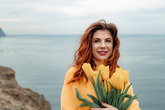 Portrait of a happy woman with hair flying in the wind against the backdrop of mountains and sea. Holding a bouquet of yellow tulips in her hands, wearing a yellow sweater.