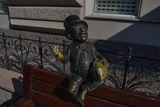 A small figurine of a man sitting on the head of a street bench. Attracts the attention of tourists.