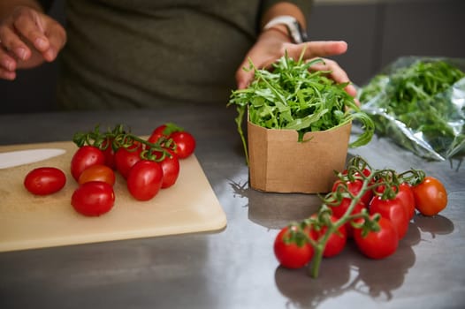 Close-up view of an unknown woman standing at kitchen counter with fresh ingredients in eco friendly recyclable cardboard bags. A bunch of red ripe tomato cherry and arugula leaves for healthy salad