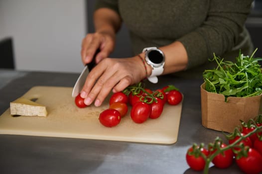 Close-up hands of a woman chopping cherry tomato on a cutting board, preparing healthy salad with fresh arugula leaves, standing at kitchen counter. Culinary, diet, healthy eating and food concept