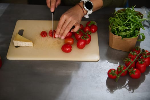 Overhead view woman chopping tomato cherry on a cutting board. A slice of delicious cheese, a bunch of organic tomatoes and fresh arugula leaves on the kitchen countertop. Italian Mediterranean food