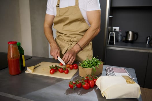 Close-up view of a male chef in beige apron, chopping red ripe tomato cherry on a cutting board. Ingredients on the kitchen table. Man cooking healthy salad in the minimalist home kitchen interior
