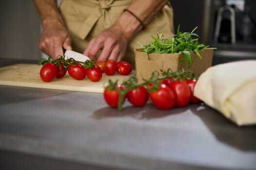 Close-up view of a male chef's hands chopping tomato cherry on a cutting board. Ingredients on the kitchen table. Man cooking healthy salad in the minimalist home kitchen interior. Food concept